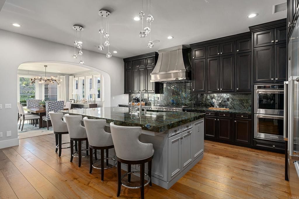 This is a well-lit kitchen featuring dark wood cabinetry, stainless steel appliances, and a large island with a green granite countertop. The island has light gray cabinets and bar stool seating. The kitchen opens to a dining area, creating an open-concept living space.
