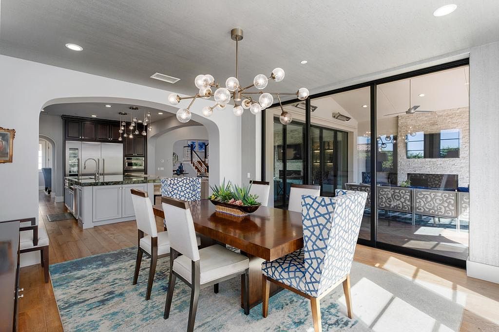 This is an interior shot of a dining room featuring a large wooden table with seating for six, including two chairs with a blue and white geometric pattern. A modern chandelier hangs above the table, and sliding glass doors lead to an outdoor patio area. The room is decorated in neutral tones with a blue and white rug, creating a sophisticated and inviting atmosphere.