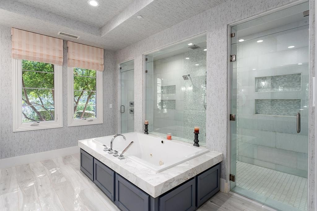This is a luxurious primary bathroom featuring a large soaking tub with a marble surround and dark blue cabinetry. Two large windows with roman shades provide natural light. There are two glass-enclosed showers, one on each side of the tub. The floor is light wood-look tile, and the walls are textured.