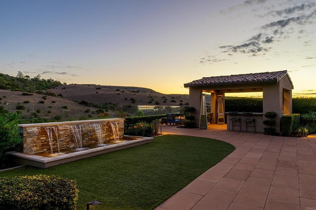 This exterior shot showcases a beautifully landscaped backyard featuring a modern water fountain, lush green lawn, and a covered outdoor bar area with seating. The patio is paved with large tiles, and the background reveals rolling hills at sunset, creating a serene and luxurious atmosphere. The perspective is from a low angle, emphasizing the depth and grandeur of the outdoor space.