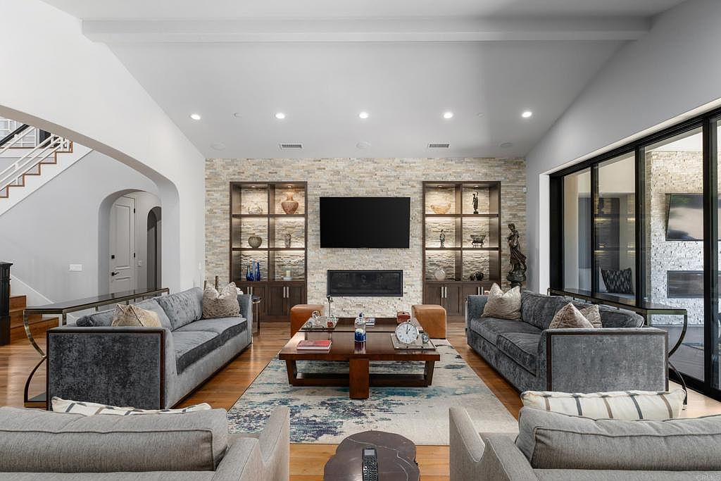 This is a spacious living room featuring two gray sofas, a wooden coffee table, and two armchairs. The focal point is a stone accent wall with a large television and built-in shelving units on either side. The room is well-lit with recessed lighting and natural light from a sliding glass door, creating a comfortable and inviting atmosphere.