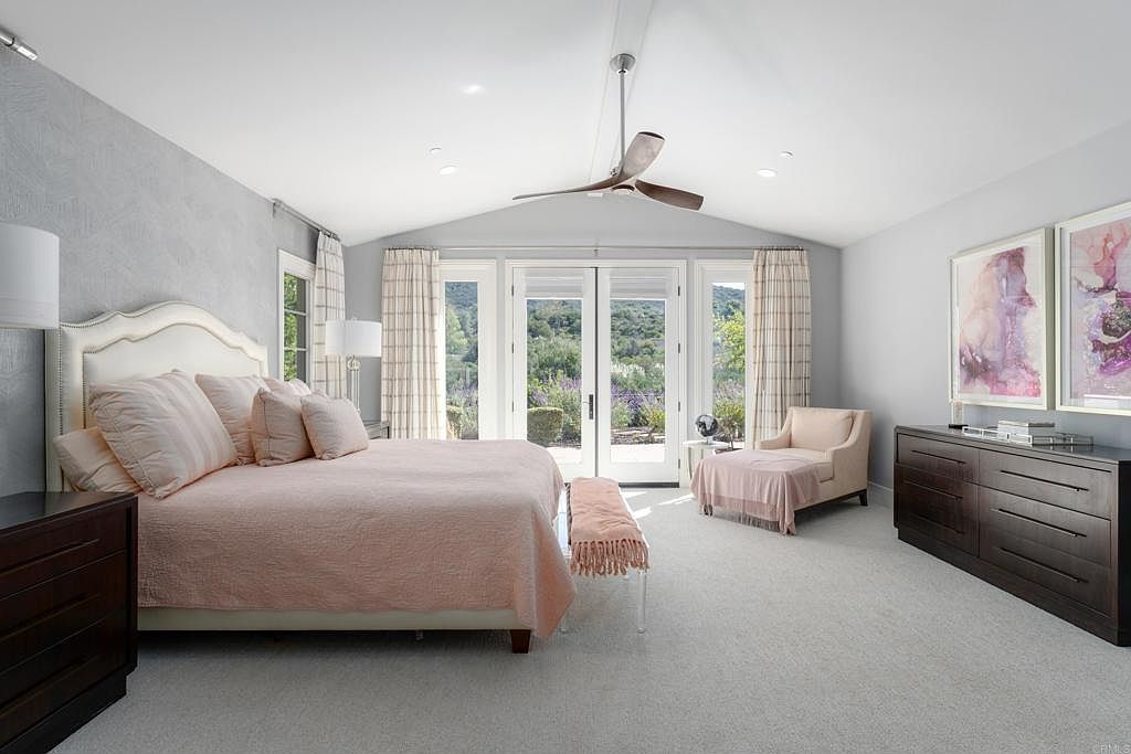 This is a bright and airy primary bedroom featuring a large bed with a light pink comforter and decorative pillows. The room has a vaulted ceiling with a ceiling fan, and large glass doors lead to an outdoor area, providing ample natural light. A dark wood dresser and artwork add a touch of elegance to the space.