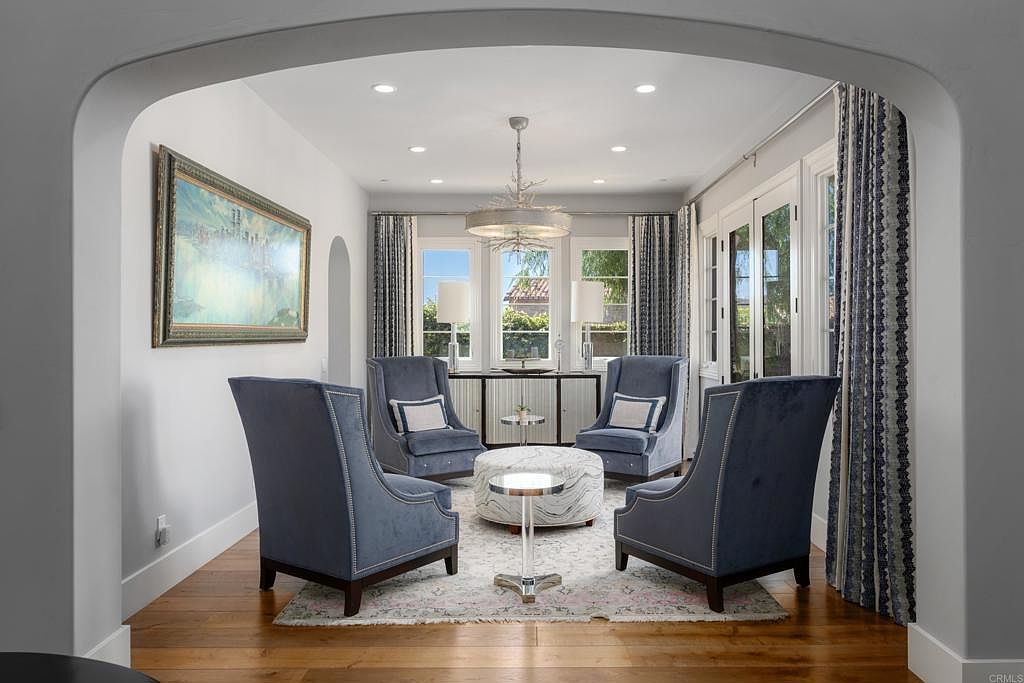 This is an interior shot of a living room featuring four blue upholstered armchairs arranged around a round ottoman and small table on a patterned rug. The room is well-lit with natural light from the windows behind the console table and an ornate chandelier. The walls are white, and a large painting hangs on the left wall, adding a touch of elegance to the space.