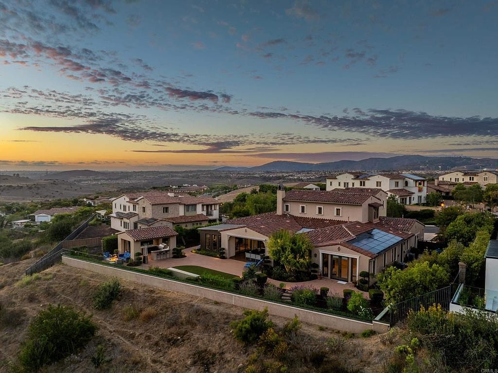 This aerial view showcases a collection of luxury homes nestled in a hillside community. The homes feature red tile roofs, stucco exteriors, and well-manicured landscaping. The image captures a serene and upscale residential setting, with a scenic backdrop of distant hills and a colorful sky at dusk.