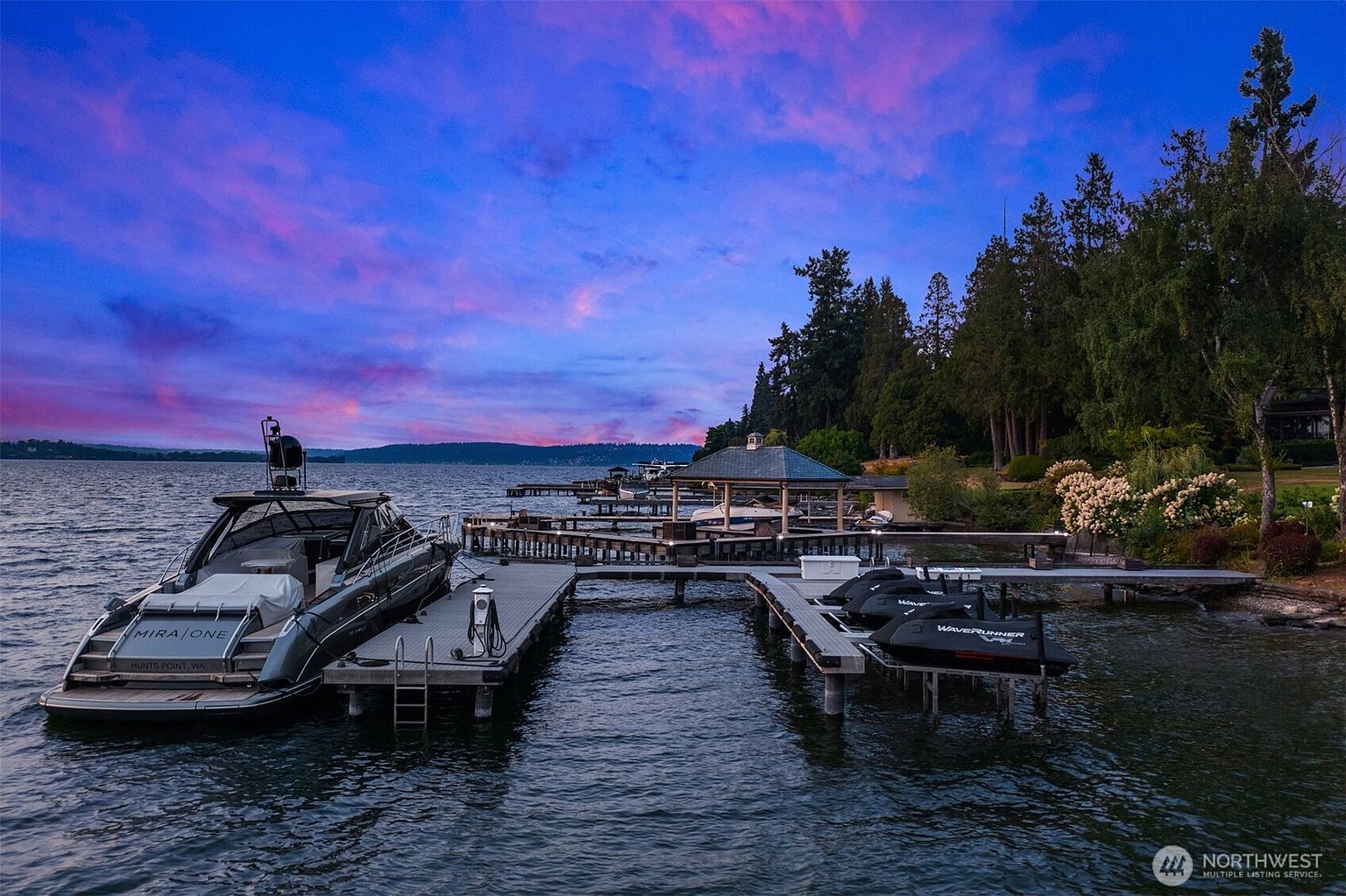 This stunning waterfront image showcases a luxurious boat docked at a private pier, accompanied by jet skis. The scene is set against a vibrant twilight sky, with lush greenery lining the shore. The overall impression is one of opulence and serene waterfront living.