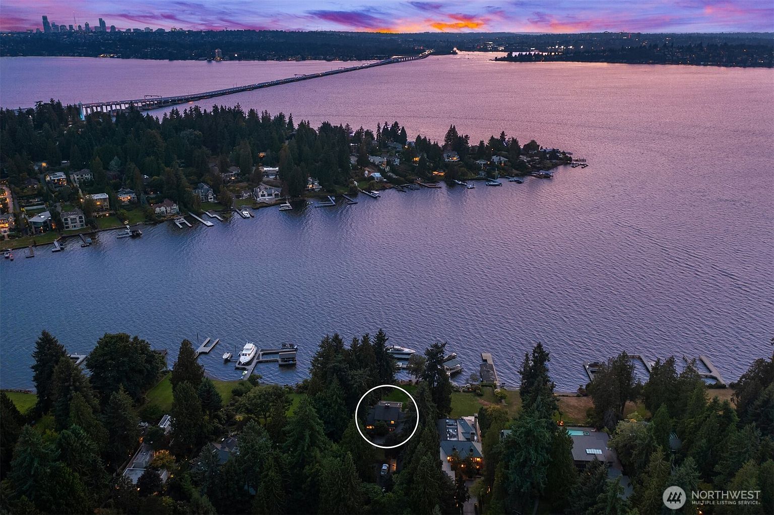 This aerial shot showcases a waterfront property nestled among lush greenery, with a private dock extending into the calm lake. The house, encircled for emphasis, is surrounded by mature trees, offering both privacy and a serene setting. In the background, a bridge stretches across the water, connecting to the distant cityscape under a vibrant sunset sky.
