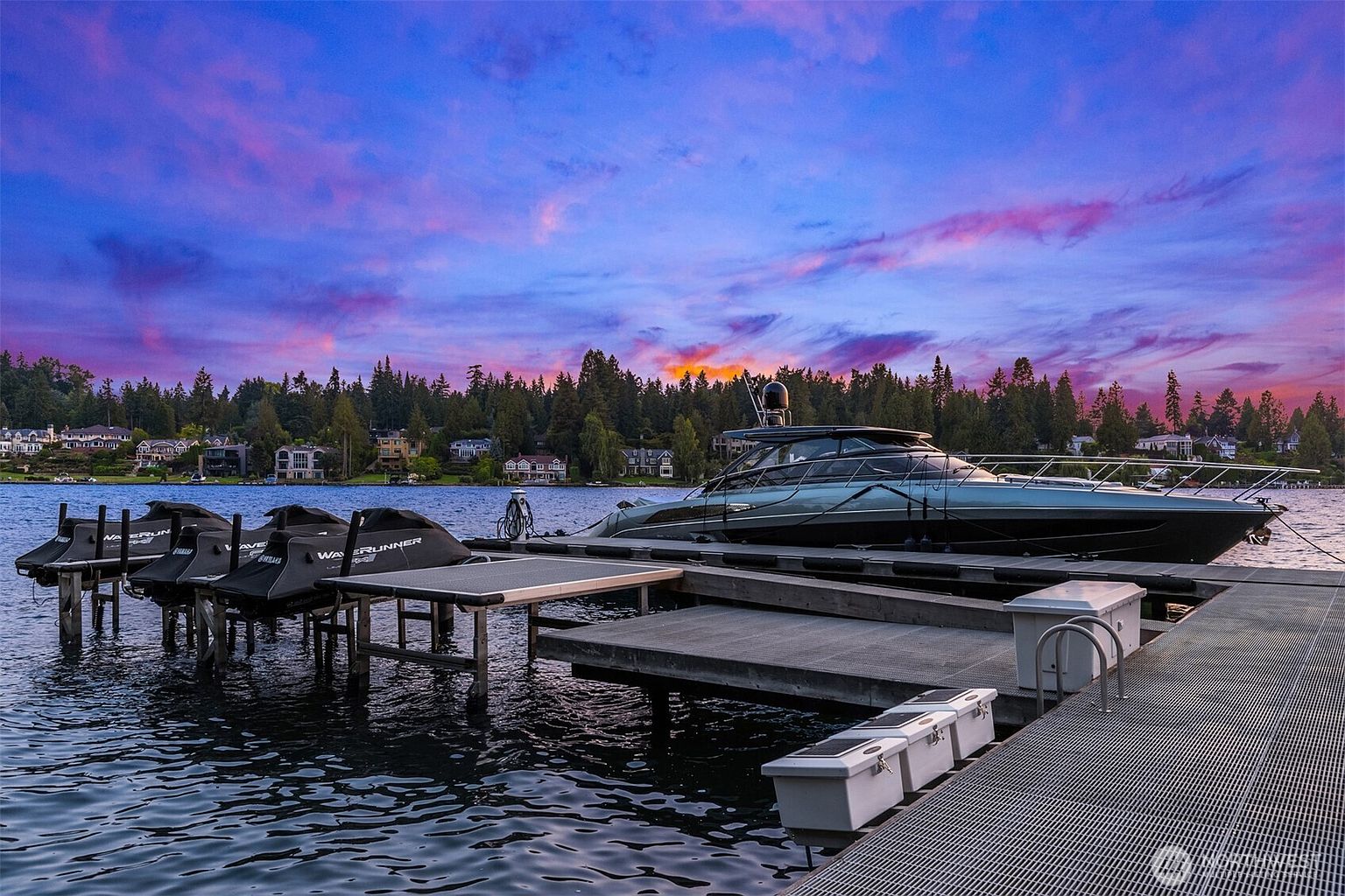 The image showcases a modern yacht docked at a well-maintained pier during a vibrant sunset. The pier features a metal grate surface and includes storage units, while the yacht exhibits sleek lines and a contemporary design. The background reveals waterfront homes nestled among lush trees, contributing to a luxurious and serene atmosphere.