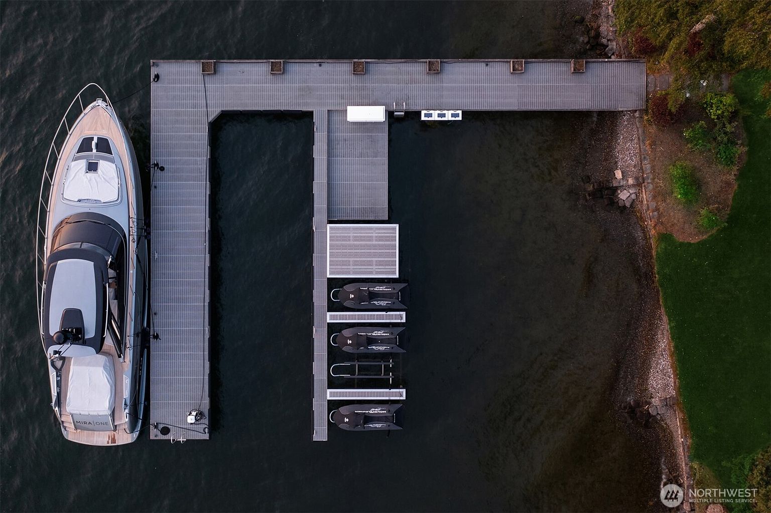 This aerial shot showcases a modern yacht docked at a well-maintained pier. The pier features a T-shaped design with multiple slips for smaller boats or jet skis. Lush greenery lines the shoreline, providing a natural contrast to the gray tones of the pier and boat.