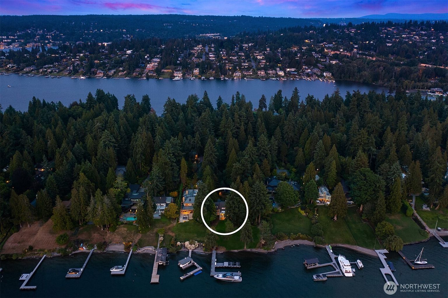This aerial shot showcases a waterfront property nestled among lush trees. The house, partially visible, sits on a well-manicured lawn leading to the water's edge, where several docks and boats are moored. The surrounding landscape includes a lake and distant residential areas, creating a serene and private setting.
