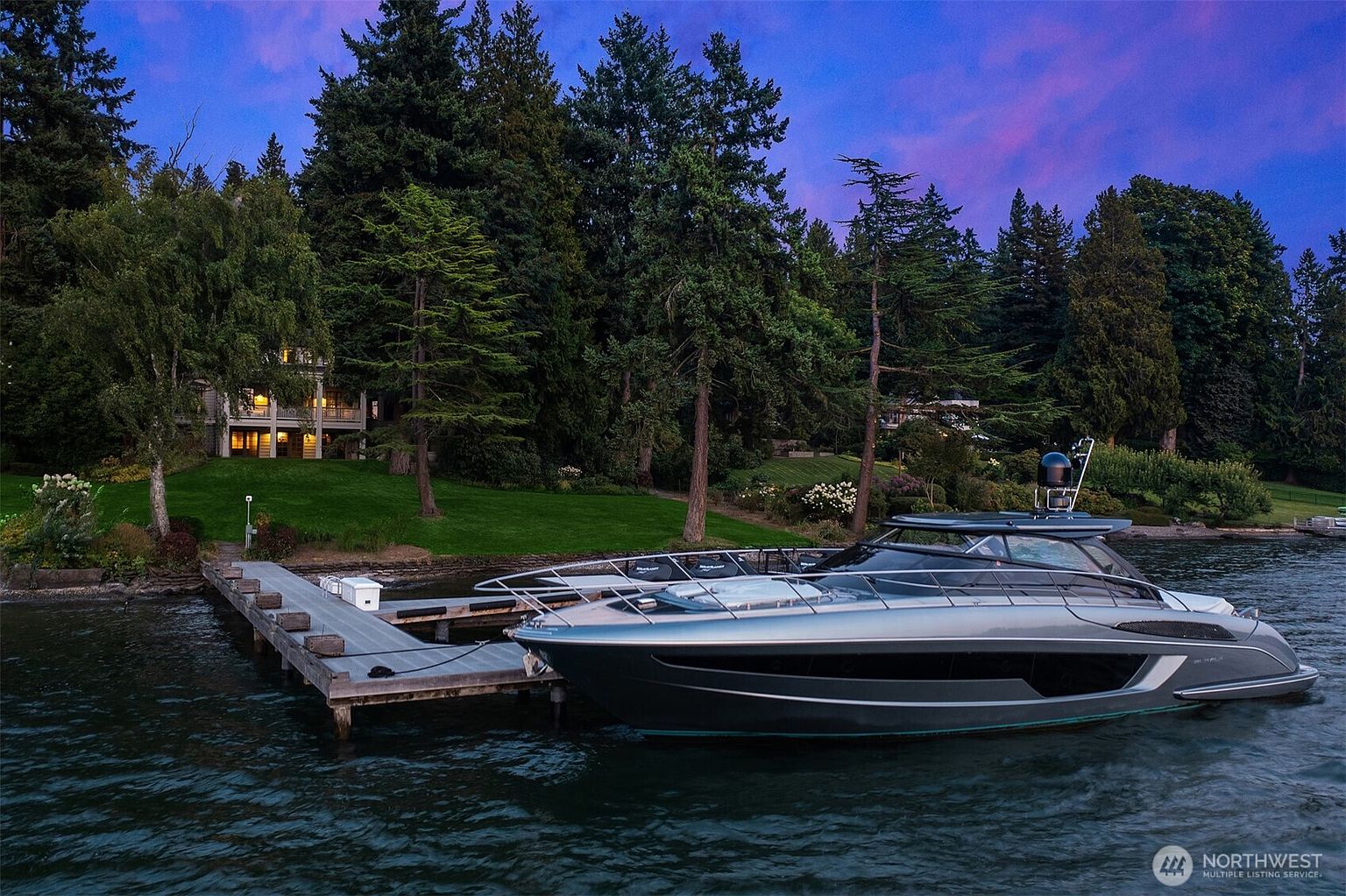 This image showcases a modern yacht docked at a private pier, with a luxurious waterfront home visible in the background. The sleek design of the yacht contrasts with the lush greenery and mature trees surrounding the property, creating an impression of upscale living and tranquility. The twilight sky adds a dramatic touch, enhancing the overall appeal.