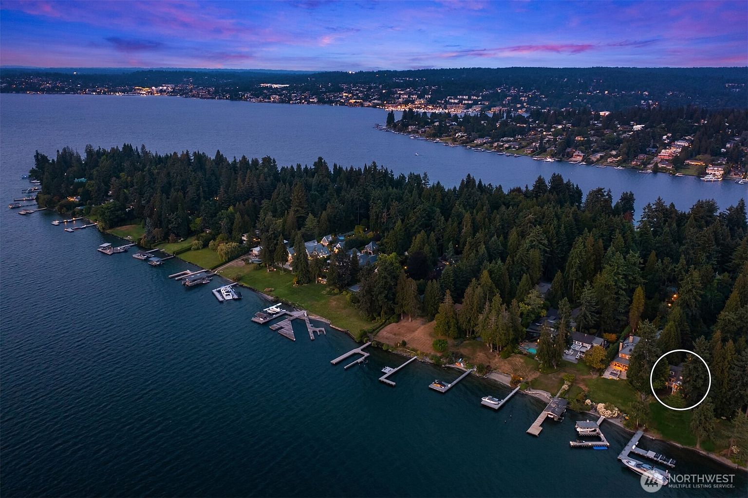 This aerial shot showcases a stunning waterfront property with lush greenery and multiple docks extending into the lake. The property features well-maintained lawns and mature trees, offering privacy and a serene atmosphere. The view extends across the lake to a distant shoreline with residential development, all under a vibrant twilight sky.