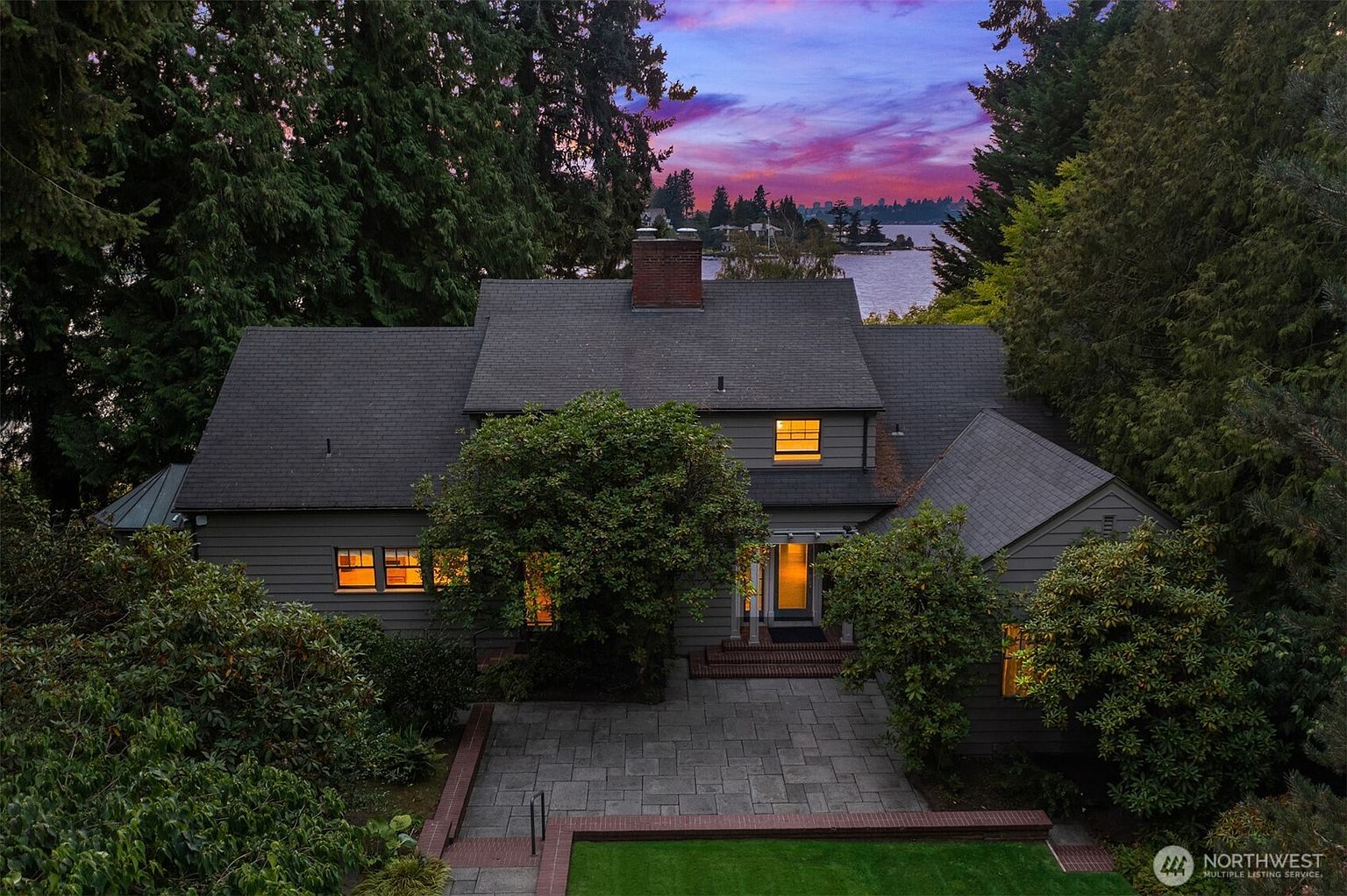 This aerial view showcases a charming home with a well-maintained gray roof, surrounded by lush greenery and mature trees. A stone patio leads to the front entrance, and the warm glow from the interior lights creates an inviting atmosphere. In the background, a serene lake and a vibrant sunset add to the property's appeal.