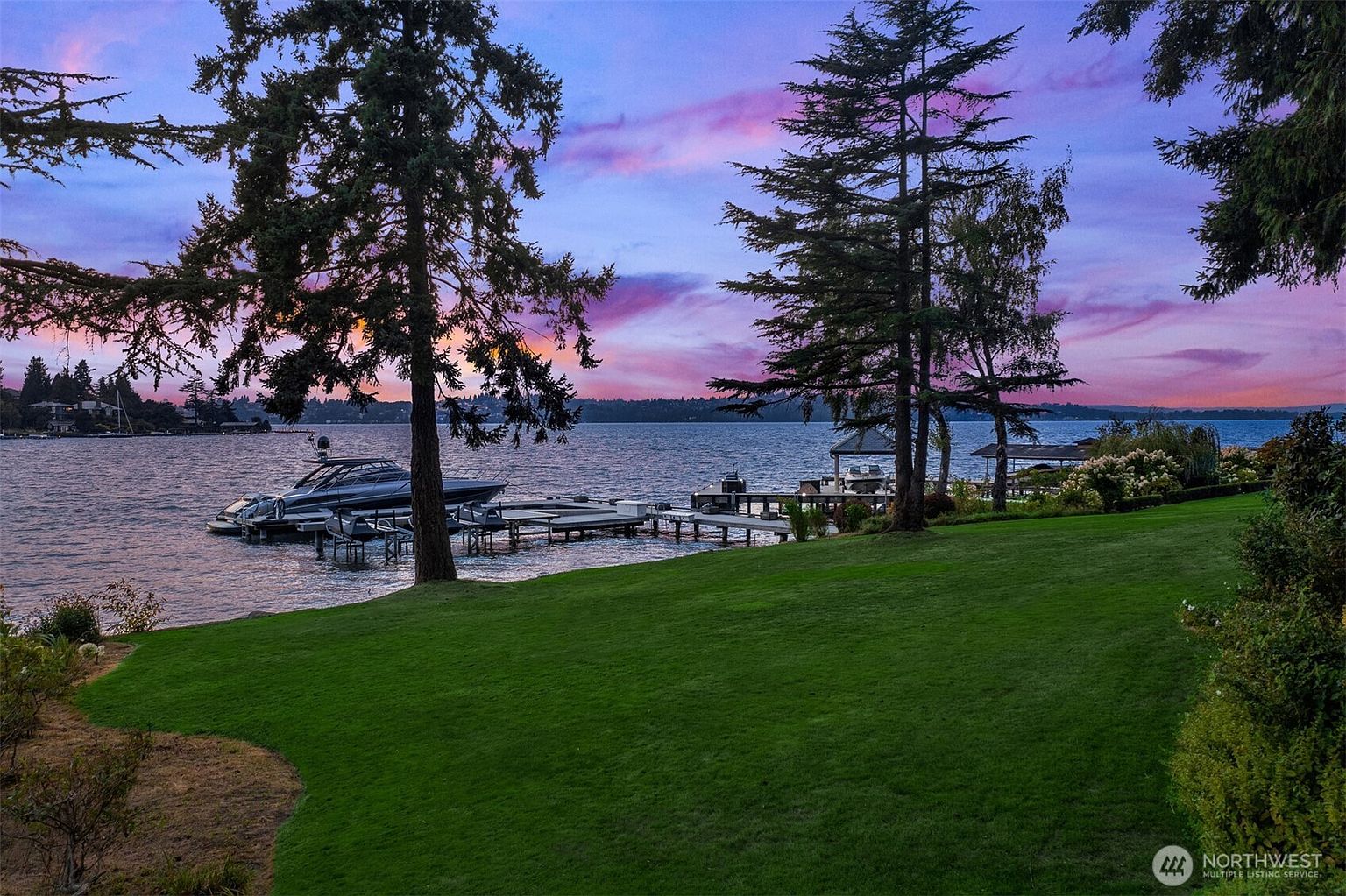 This image showcases a beautiful waterfront property with a lush green lawn extending to the water's edge. A private dock with boats is visible, and mature trees frame the scene, creating a serene and picturesque setting. The vibrant sunset adds a touch of elegance and tranquility, making it an ideal location for relaxation and recreation.