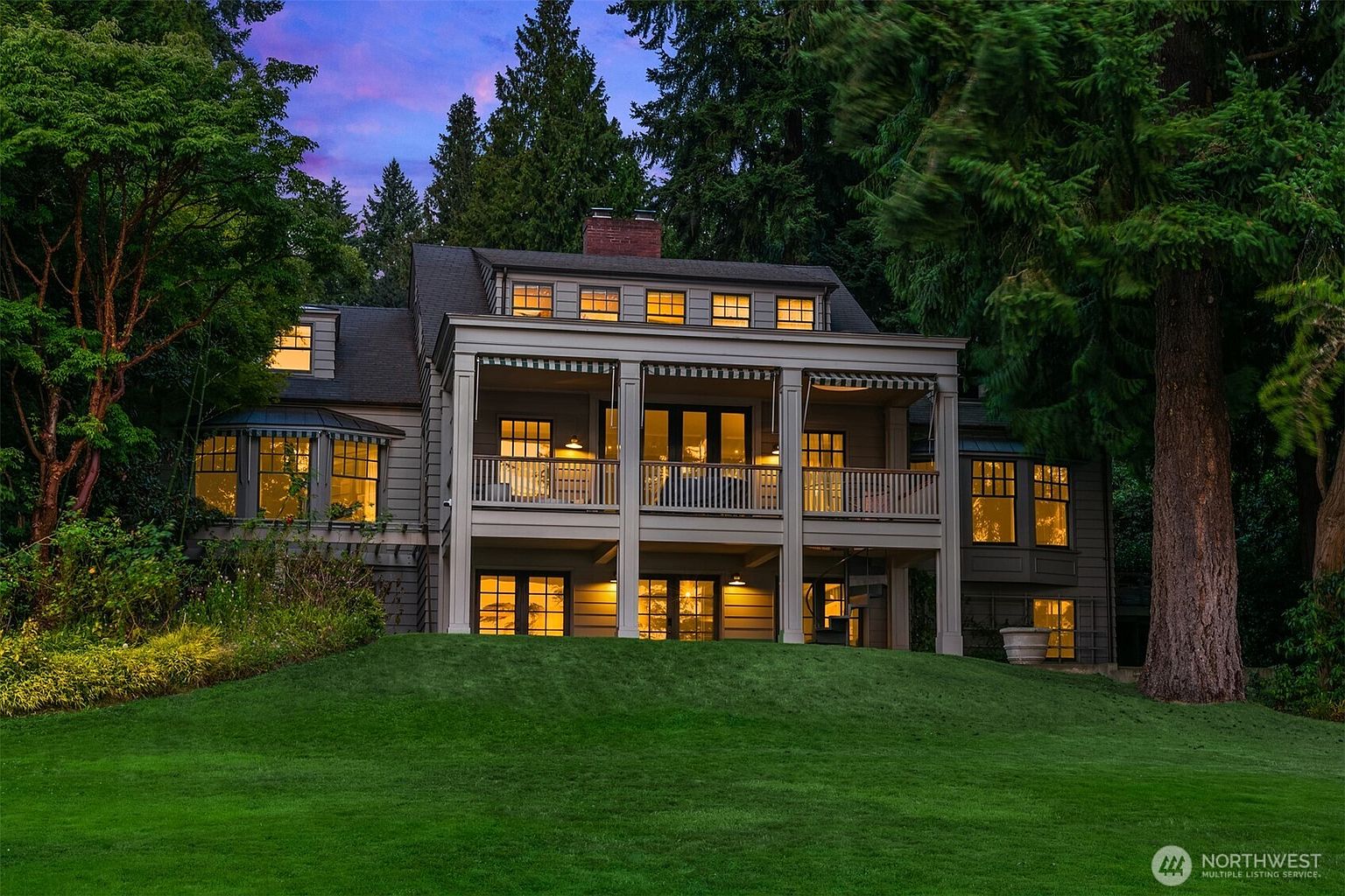 The image showcases the rear exterior of a multi-story home with a covered balcony and a well-manicured lawn. Warm light emanates from the windows, creating an inviting ambiance against the backdrop of mature trees and a twilight sky. The architecture features a classic design with a modern touch, emphasizing outdoor living and a connection to nature.