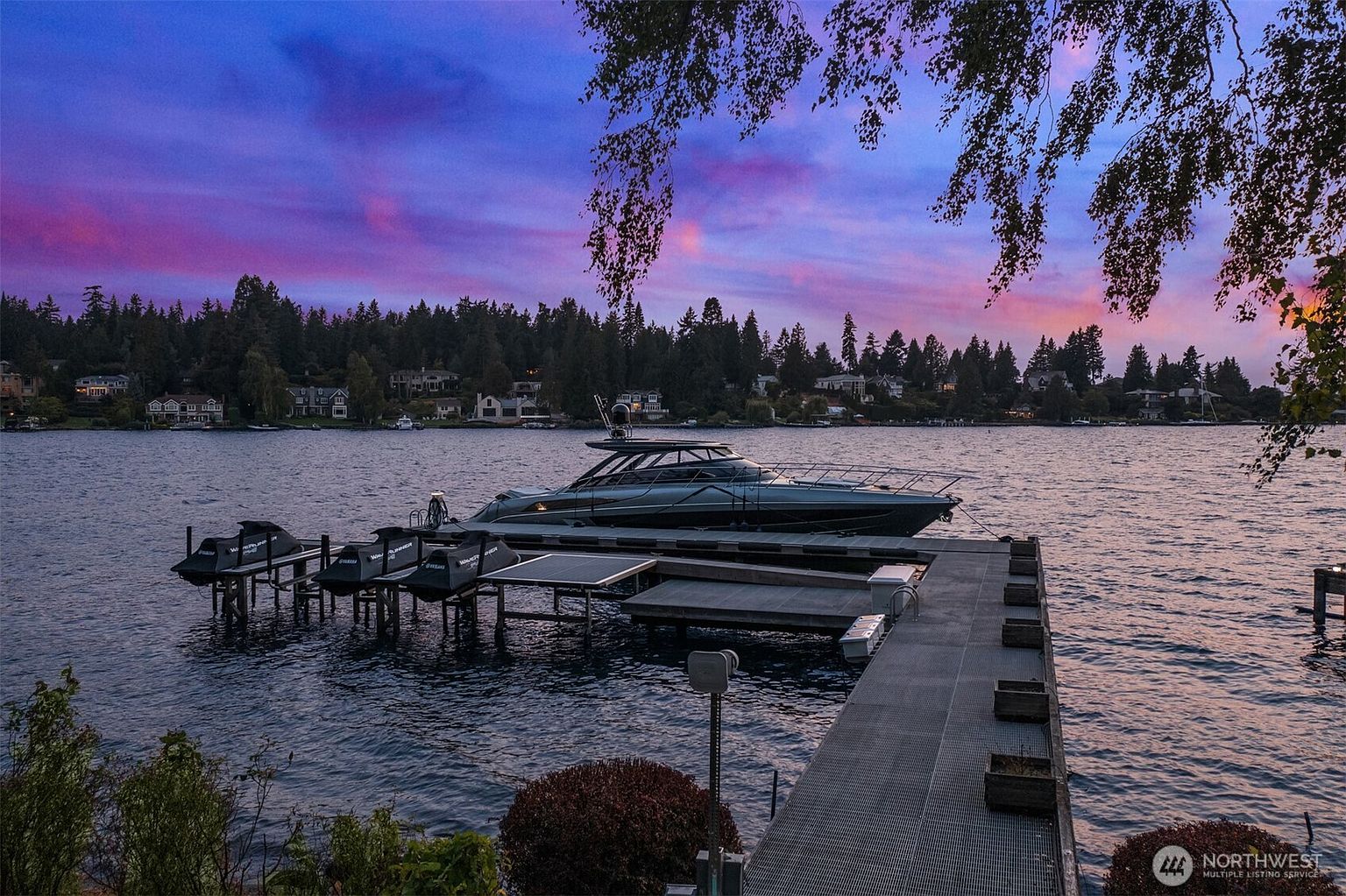 This stunning rear view showcases a luxurious waterfront property with a private dock. A sleek yacht is moored alongside the dock, accompanied by several jet skis. The backdrop features a serene lake and a picturesque treeline under a vibrant sunset sky, creating an idyllic and exclusive ambiance.