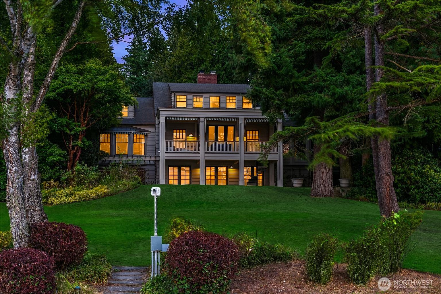 This image showcases the rear exterior of a two-story home with a covered balcony on the second floor. The house is surrounded by lush greenery and trees, creating a private and serene setting. The well-maintained lawn slopes gently towards the camera, enhancing the property's curb appeal.