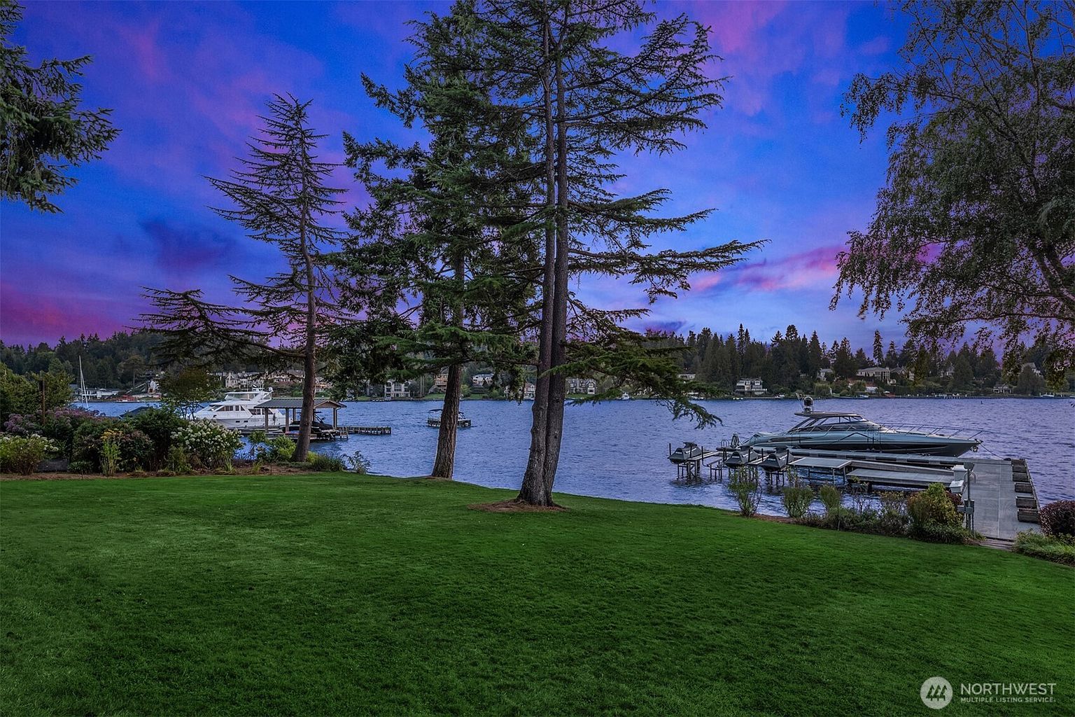 This image showcases a beautifully maintained yard with lush green grass leading to a waterfront view. Several boats are docked along the shoreline, and mature trees frame the scene. The sky displays a vibrant mix of blue and purple hues, creating a serene and picturesque setting.