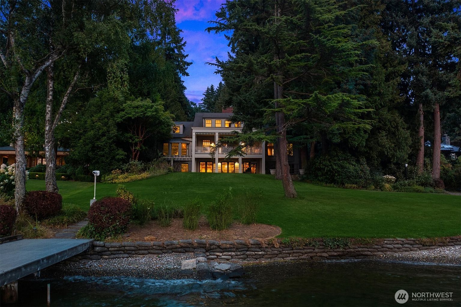 This image showcases the rear exterior of a multi-story home, beautifully situated on a lush green lawn that slopes down to the water's edge. The house features multiple balconies and large windows, suggesting ample natural light and scenic views. Mature trees frame the property, creating a sense of privacy and tranquility.