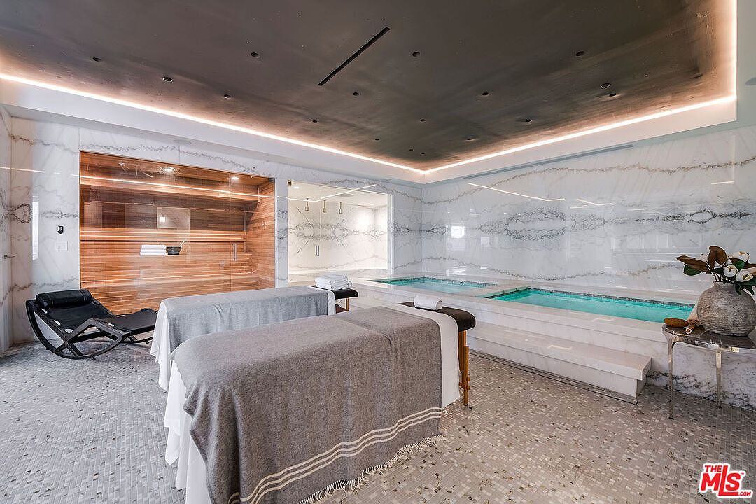 This interior shot showcases a luxurious home gym featuring two massage tables, a sauna, and a small indoor pool. The walls are adorned with marble, and the ceiling has recessed lighting, creating a spa-like atmosphere. The perspective is from the entrance, capturing the full scope of the room's amenities.