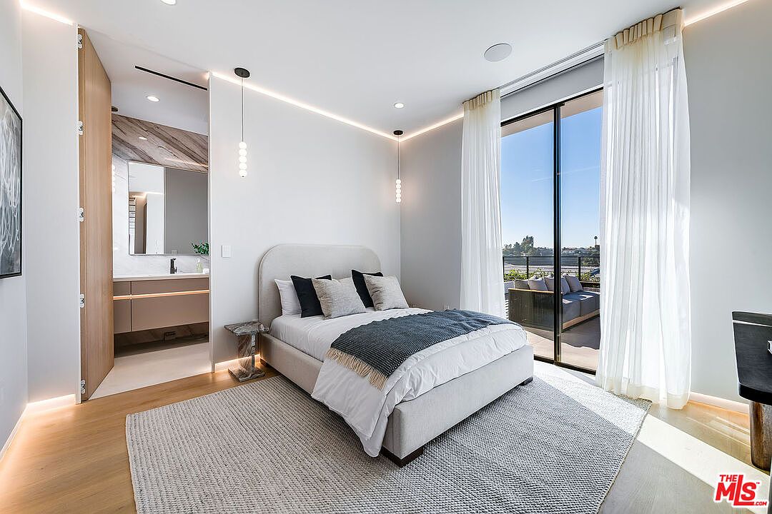 This is a bright and modern primary bedroom featuring a neutral color palette with a large bed, stylish lighting fixtures, and access to an outdoor balcony through sliding glass doors. The room is well-lit with natural light and features a contemporary design with clean lines and minimalist decor, creating a serene and inviting atmosphere. The camera is positioned at a medium angle, capturing the bed and balcony in a wide shot.