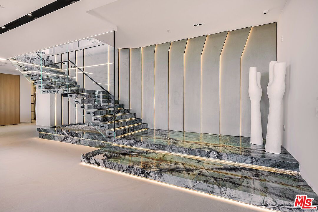 This interior shot showcases a modern hallway and staircase with luxurious design elements. The staircase features marble steps with integrated lighting, glass railings, and black metal accents. A textured wall with vertical lighting strips and two white sculptures add to the contemporary aesthetic, creating a sophisticated and elegant entryway.