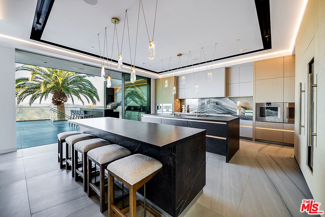 This is a modern, high-end kitchen featuring a large island with seating, sleek cabinetry, and pendant lighting. The kitchen opens to an outdoor area with a palm tree, creating a seamless indoor-outdoor living experience. The overall impression is luxurious and contemporary.