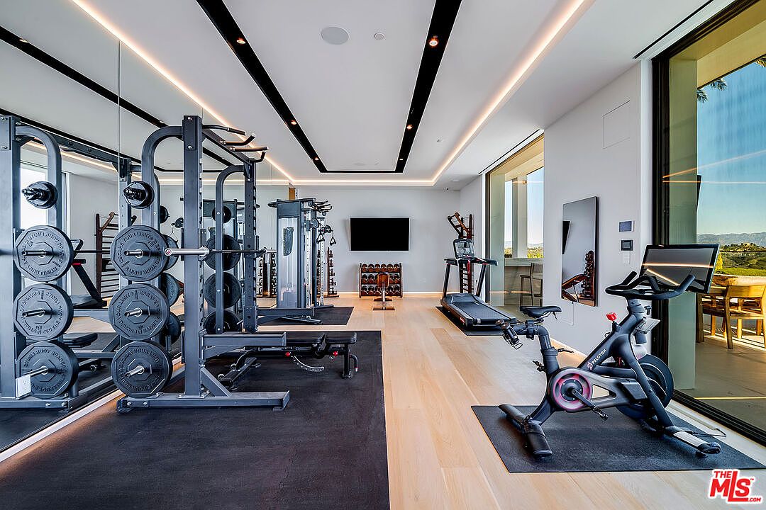 This is a well-equipped home gym featuring weightlifting equipment, a treadmill, and a stationary bike. The room has a modern design with black accents, light wood flooring, and large windows offering natural light and views. The space is designed for a comprehensive workout experience.