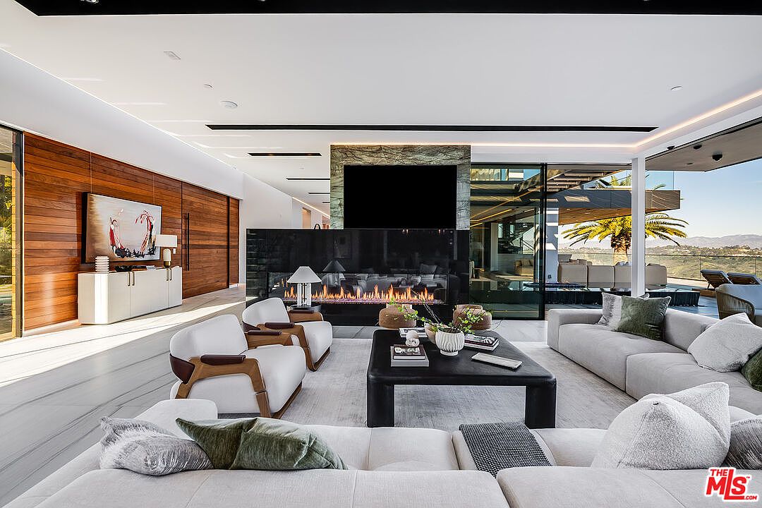 This is a luxurious living room featuring a modern fireplace with a large television above it. The room is furnished with comfortable sofas and chairs, and a sleek coffee table sits in the center. Large windows offer stunning views of the surrounding landscape, creating a bright and airy atmosphere.