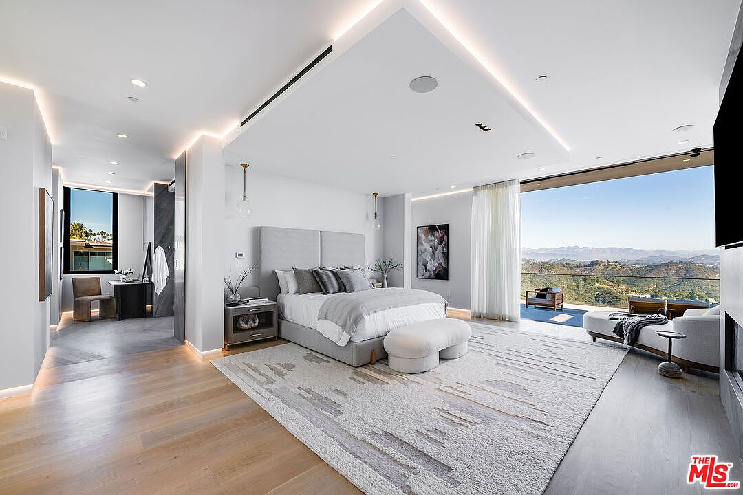 This is a luxurious primary bedroom featuring a large bed with a padded headboard, a neutral-toned rug, and floor-to-ceiling windows offering stunning mountain views. The room is decorated in a modern style with recessed lighting and minimalist decor. The open layout leads to an adjacent bathroom and a seating area on the balcony, creating a seamless indoor-outdoor living experience.