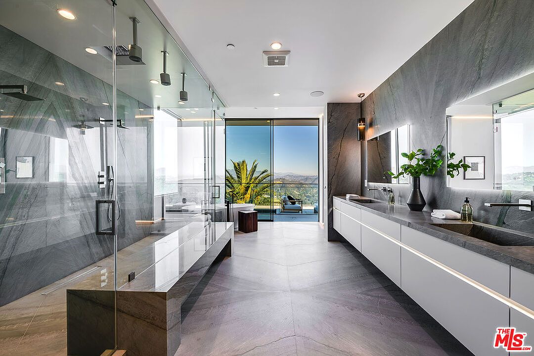 This is a luxurious primary bathroom featuring a spacious walk-in shower with glass enclosure and dual shower heads, a long floating vanity with dual sinks, and a sliding glass door leading to a balcony with scenic views. The bathroom is finished with gray marble walls and flooring, creating a sleek and modern aesthetic. The perspective is from the entrance of the bathroom, showcasing the length of the room and the view outside.