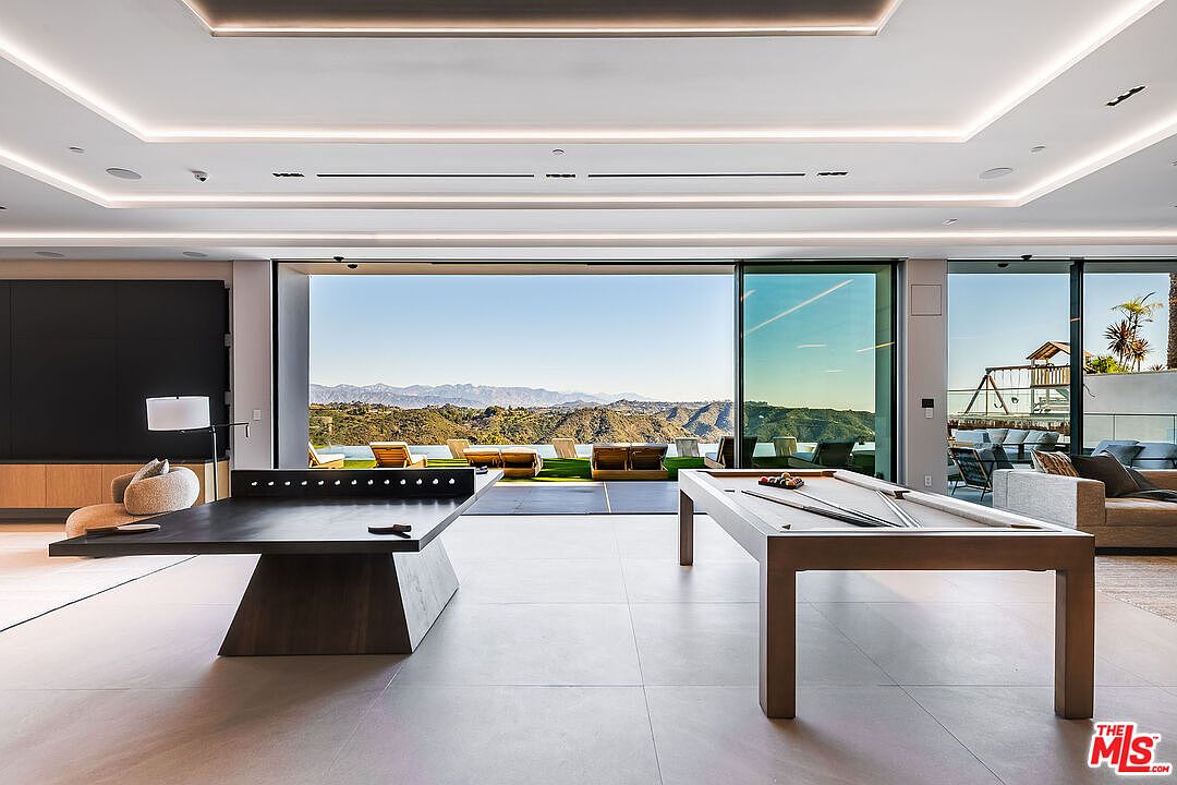 This interior shot showcases a spacious living room with a modern design, featuring a ping pong table and a pool table. Large sliding glass doors offer panoramic views of the landscape, seamlessly blending indoor and outdoor living. The room is well-lit with recessed lighting and natural light, creating an inviting and luxurious atmosphere.