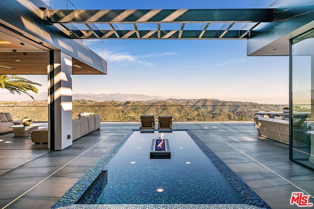 This image showcases a luxurious outdoor pool area with a stunning view of rolling hills and a clear blue sky. The pool features a unique fire pit element in the center, surrounded by elegant tiling and sleek, modern architecture. The space is designed for relaxation and entertainment, blending indoor and outdoor living seamlessly.