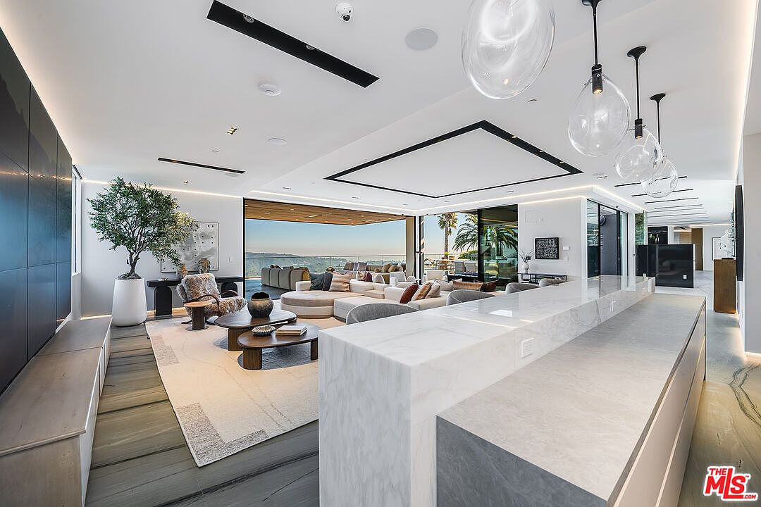 This is a luxurious living room featuring an open floor plan with expansive views of the outdoors. The room is furnished with modern, comfortable seating arrangements, including a large sectional sofa and stylish armchairs. The space is well-lit with natural light and contemporary pendant lighting, creating a bright and inviting atmosphere.