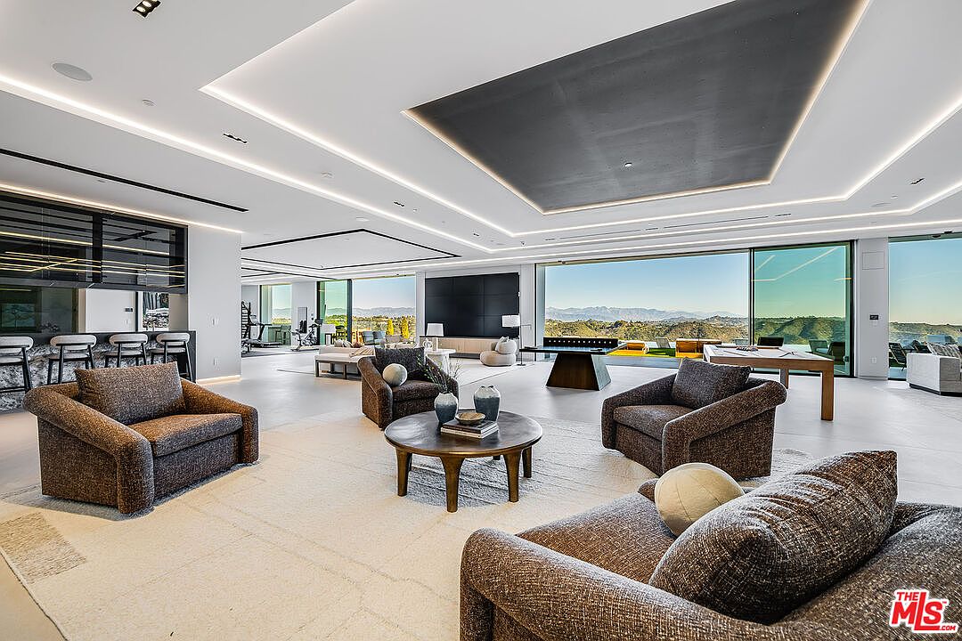 This expansive living room features a modern design with neutral tones and abundant natural light. The space includes multiple seating areas with plush armchairs and sofas, centered around a round coffee table. Large windows offer panoramic views of the surrounding landscape, enhancing the luxurious and open feel of the room.