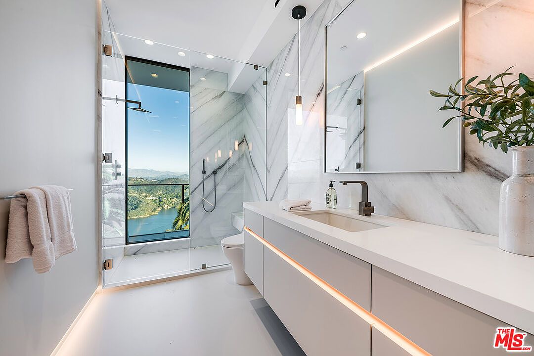 This is a luxurious primary bathroom featuring marble walls and a glass-enclosed shower with a stunning view. The vanity has a sleek, modern design with integrated lighting, and the overall aesthetic is clean and contemporary. The space is well-lit and appears spacious, creating a spa-like atmosphere.