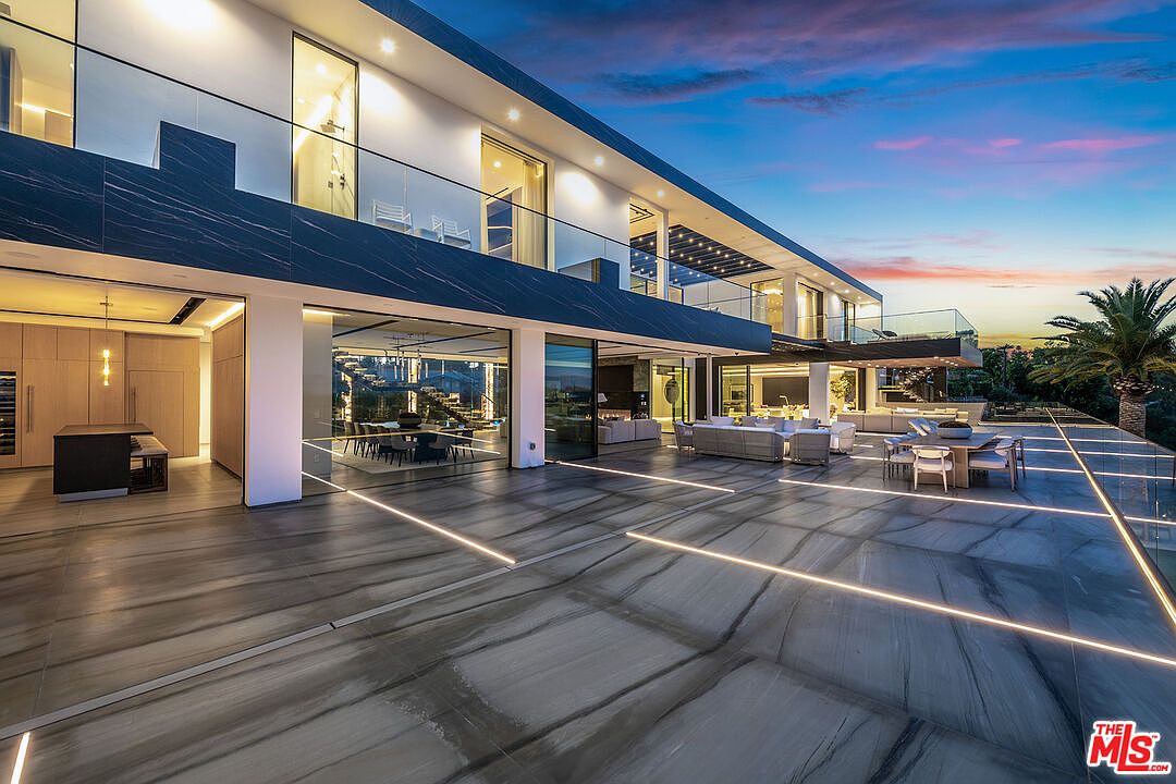 This image showcases a luxurious patio/deck area of a modern home at dusk. The expansive outdoor space features sleek, gray marble flooring with integrated linear lighting, creating a sophisticated ambiance. Outdoor seating arrangements and glass railings offer unobstructed views, while the open floor plan seamlessly connects indoor and outdoor living spaces, enhancing the property's appeal.