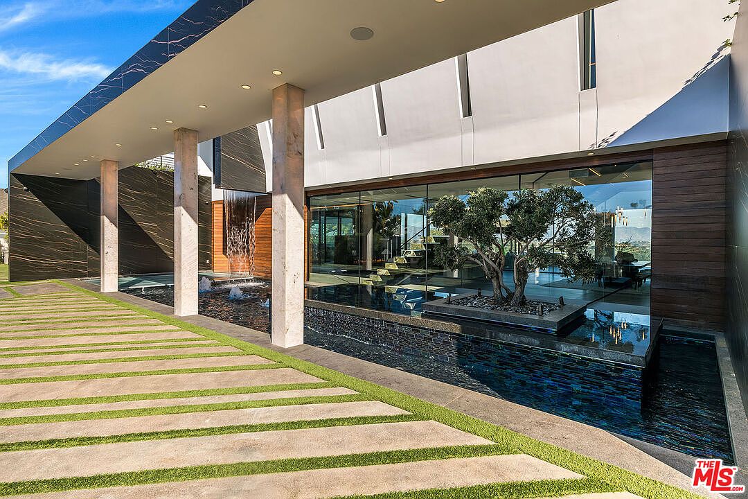 This image showcases the grand entryway of a modern home, featuring a striking combination of architectural elements. A covered walkway with marble columns leads to a glass-walled entrance, reflecting a serene water feature with a mature tree. The walkway is paved with concrete slabs interspersed with green grass, creating a visually appealing and inviting approach.
