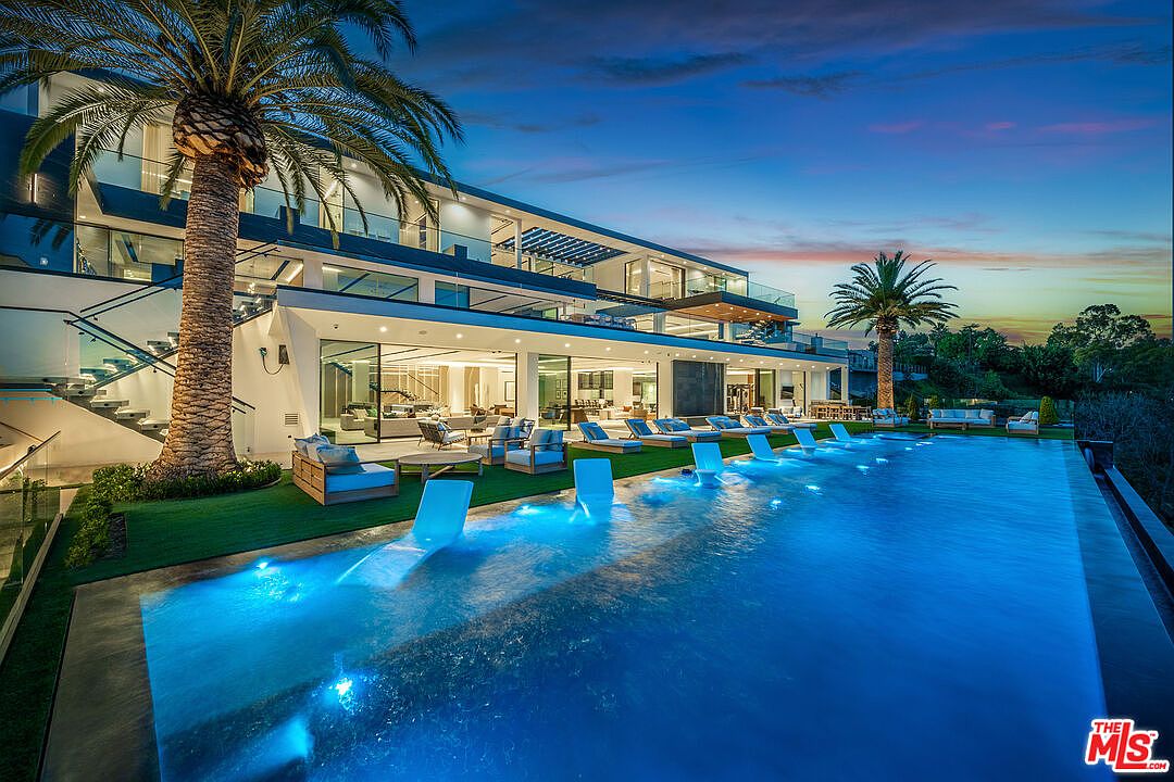 This image showcases a luxurious infinity pool at dusk, featuring built-in lounge chairs with underwater lighting. The pool is adjacent to a modern, multi-story home with expansive glass walls and balconies, surrounded by palm trees and lush landscaping. The scene evokes a sense of high-end living and relaxation.