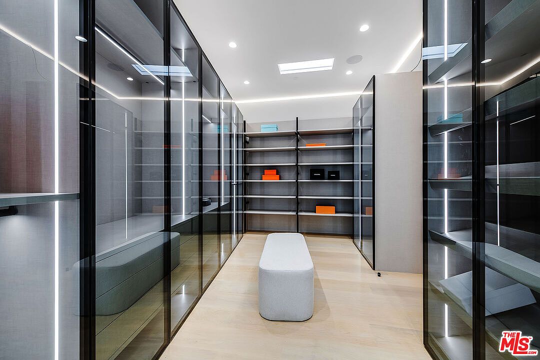 This is a modern walk-in closet featuring glass-fronted wardrobes with integrated lighting and open shelving. A central ottoman provides a seating area, and the light wood flooring adds warmth to the space. The overall impression is one of luxury and organization.