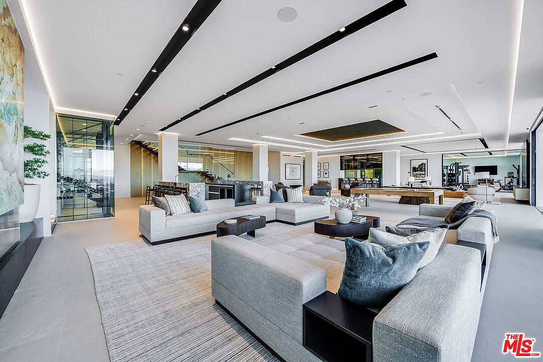 This is a spacious and modern living room featuring a large sectional sofa with plush pillows, a stylish coffee table, and a neutral-toned rug. The room has a sleek, minimalist design with black ceiling accents and recessed lighting. In the background, there is a wine cellar, a bar area, a pool table, and a home gym, creating a luxurious and multi-functional living space.