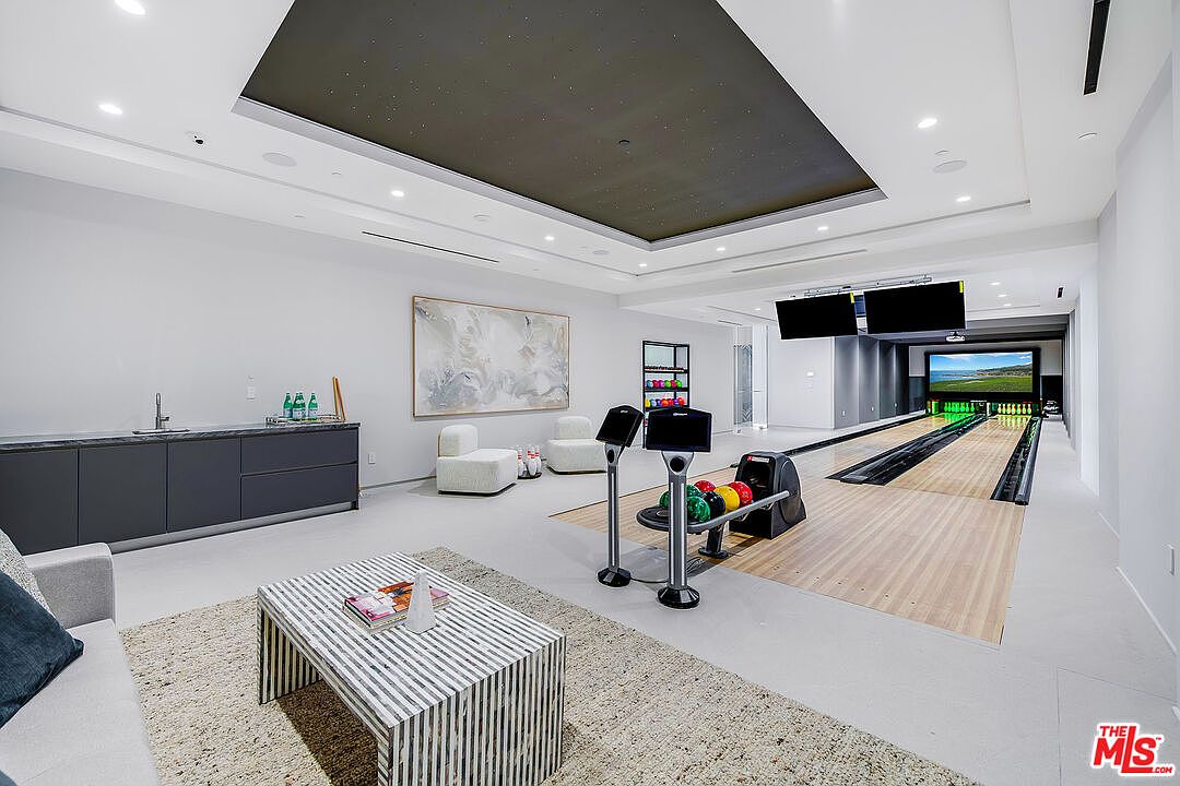 This interior shot showcases a luxurious bowling alley within a home. The space features a modern design with a dark ceiling accent, sleek furniture, and a wet bar area. The bowling lanes are well-lit, and there's a large screen at the end of the lanes, creating an entertainment-focused atmosphere.