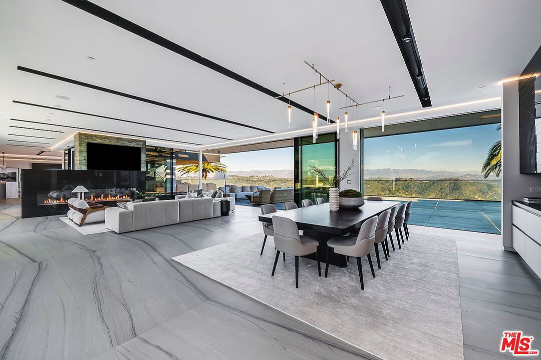 This interior shot showcases a luxurious dining area within an open-concept living space. A long, dark wood dining table is surrounded by modern gray chairs, set upon a light-colored rug. Floor-to-ceiling windows offer panoramic views of the landscape, while sleek black ceiling beams and contemporary lighting fixtures add to the sophisticated ambiance.