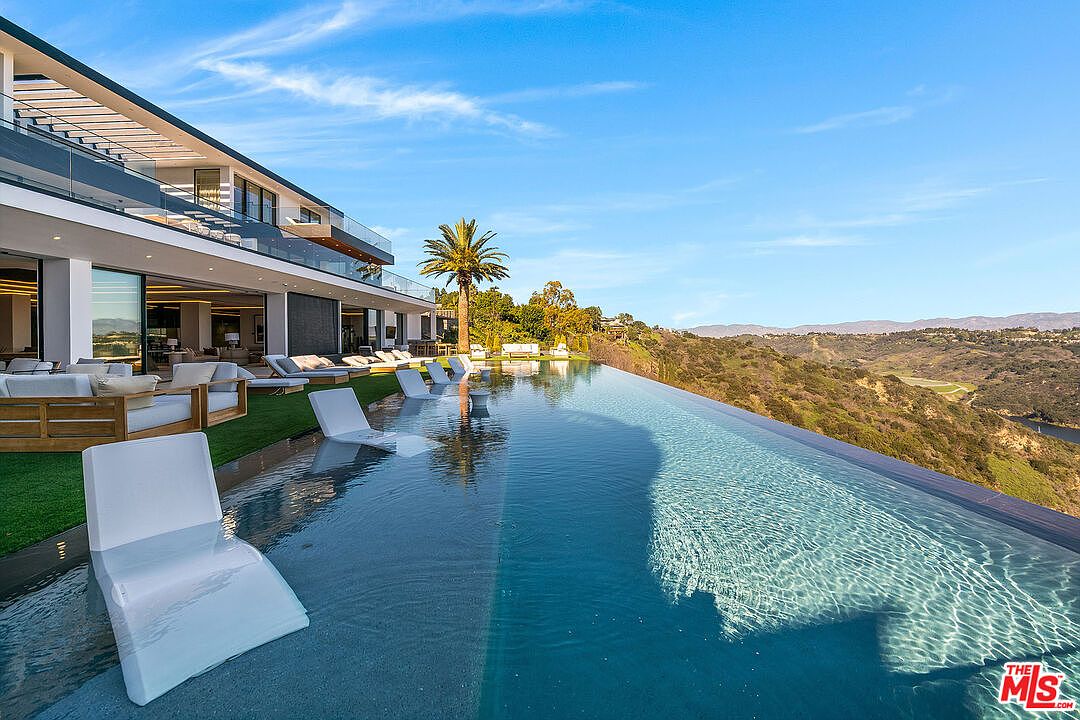 This image showcases a luxurious infinity pool extending towards a scenic vista of rolling hills and a clear blue sky. The pool is surrounded by modern lounge chairs and is adjacent to a contemporary home with expansive glass doors, creating a seamless indoor-outdoor living experience. The overall impression is one of opulence and tranquility.