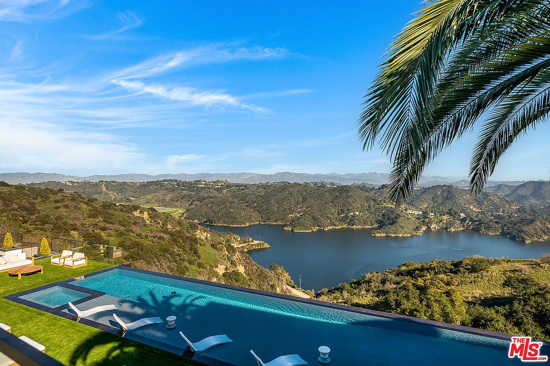 This image showcases a luxurious infinity pool overlooking a stunning landscape with a lake and rolling hills. The pool area features modern lounge chairs and a spa, creating an inviting outdoor space. A palm tree adds a tropical touch, enhancing the overall sense of relaxation and exclusivity.
