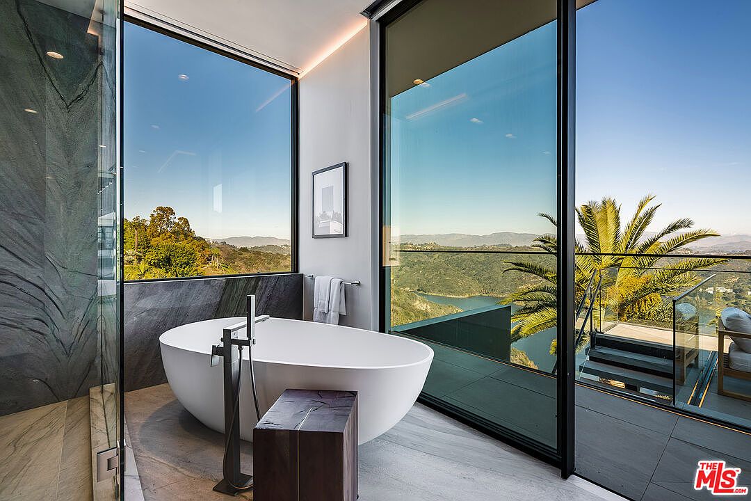 This is a luxurious primary bathroom featuring a freestanding white bathtub positioned in front of a large window with stunning views of the landscape. The bathroom has a modern design with gray marble accents, a dark wood side table, and sleek black fixtures. The open layout and natural light create a spa-like atmosphere.