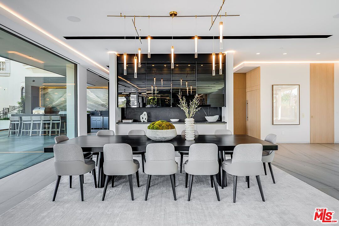 This is a modern dining room featuring a long, dark wood table surrounded by light gray upholstered chairs with black legs. A striking linear chandelier hangs above the table, and a large decorative centerpiece sits in the middle. The room has a clean, minimalist aesthetic with light wood accents and a large sliding glass door leading to an outdoor area.