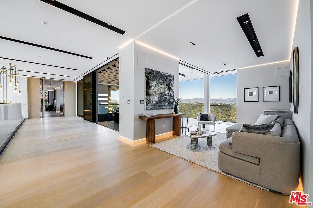 This is a spacious and modern living room featuring light wood flooring, recessed lighting, and large windows offering scenic views. The room includes a comfortable sofa, a stylish coffee table, and a console table with artwork above it. The open layout and minimalist design create a luxurious and inviting atmosphere.