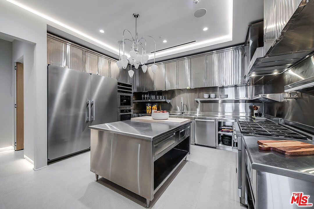 This is a modern, professional-grade kitchen featuring stainless steel cabinetry, countertops, and appliances, including a large refrigerator and a commercial-style range. A central island provides additional workspace, and a unique chandelier hangs above. The overall impression is sleek, functional, and high-end.