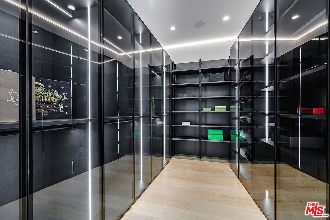 This is a modern walk-in closet featuring dark shelving and glass-fronted cabinets with integrated lighting. The closet has a light wood floor and is well-organized with boxes and accessories on display. The perspective is from the entrance of the closet, showcasing the depth and storage capacity.