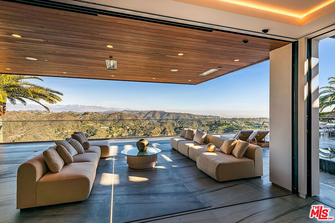 This image showcases a luxurious outdoor living space with stunning panoramic views of rolling hills and distant mountains. The patio features two plush, neutral-toned sofas arranged around a modern glass coffee table, all under a sleek wooden ceiling with recessed lighting. The open design seamlessly blends indoor and outdoor living, creating an inviting and sophisticated atmosphere perfect for relaxation and entertainment.
